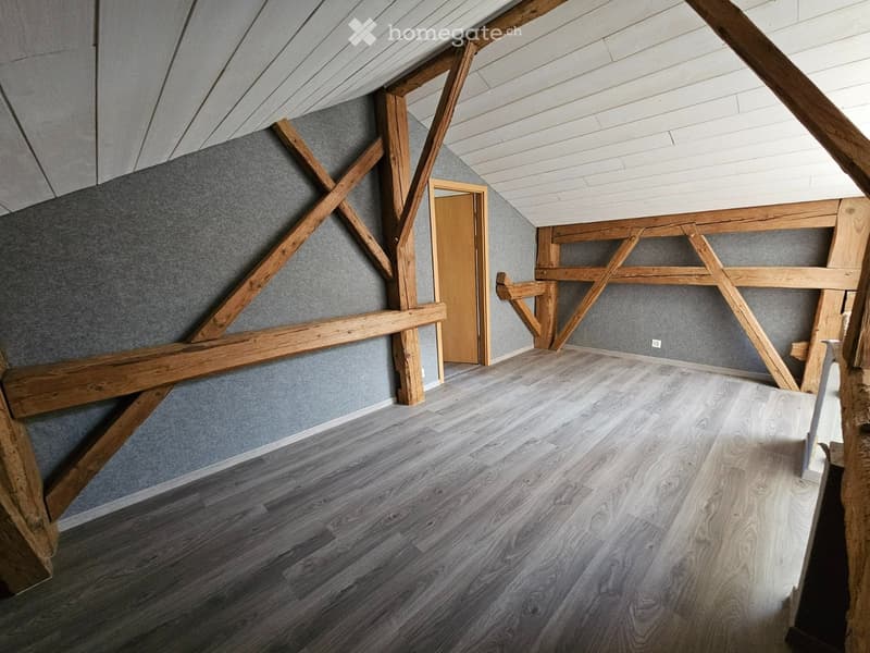 Empty attic space with wooden beams, grey walls, and a wooden door leading to another area.
