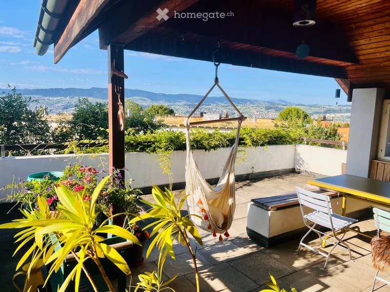 Balcony with a hammock, table, chairs, and potted plants, overlooking a scenic valley and mountains.