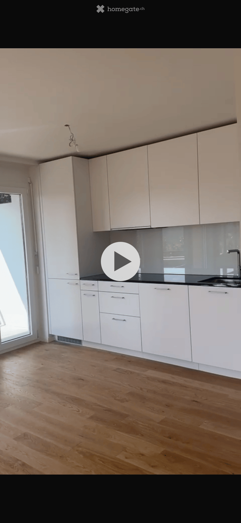 Empty kitchen with white cabinets, wooden floor, and a large window letting in natural light.