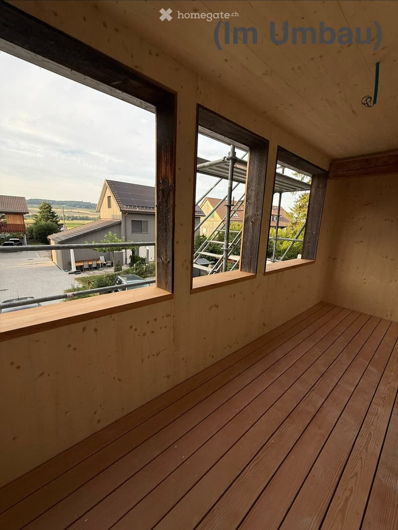 Wooden balcony with wooden decking, metal railing, and multiple windows overlooking a rural landscape with houses and fields.