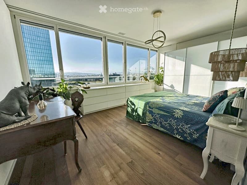 Bright bedroom with large windows offering a city view, featuring a bed with a patterned duvet, a nightstand with a lamp, a wooden table with a decorative item, and a hanging woven lamp.