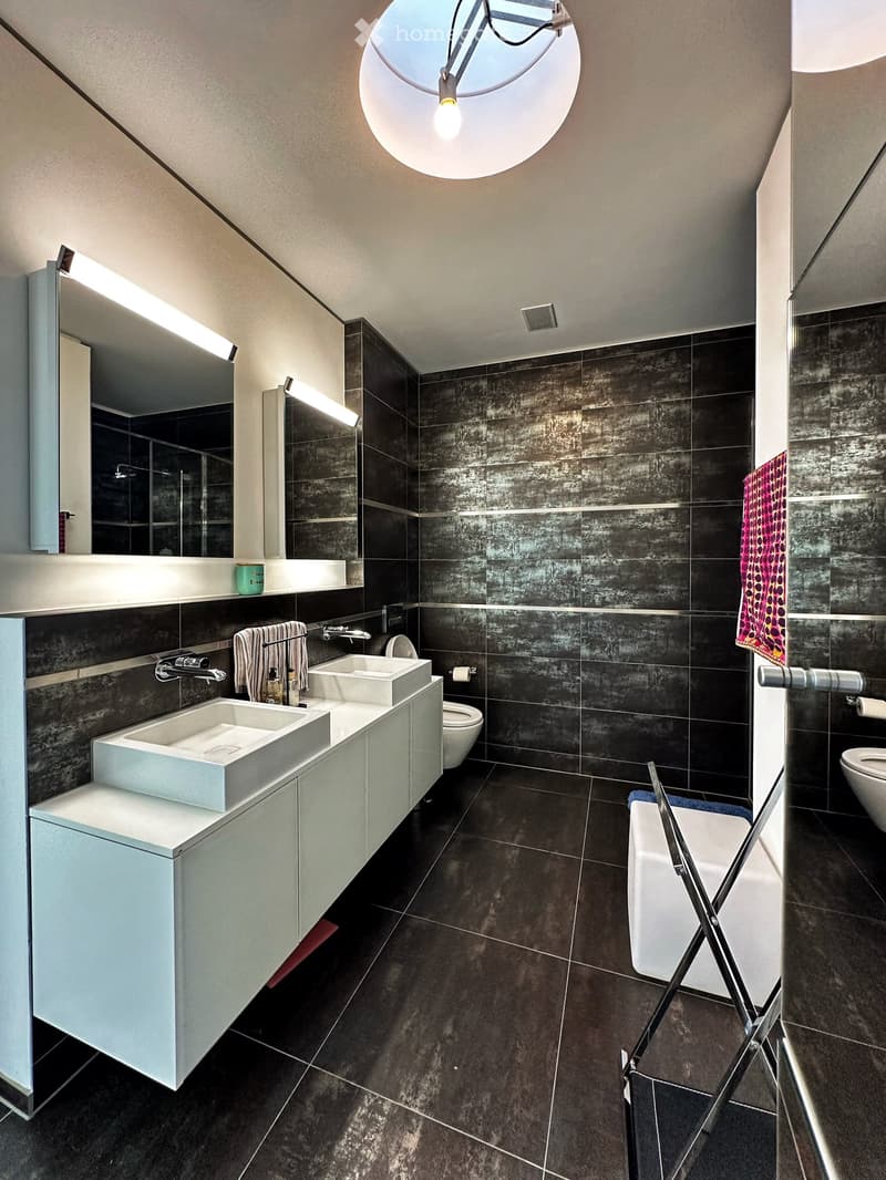 Modern bathroom with dark tiled walls and floor, white sink and cabinet, large mirror, and two sinks.