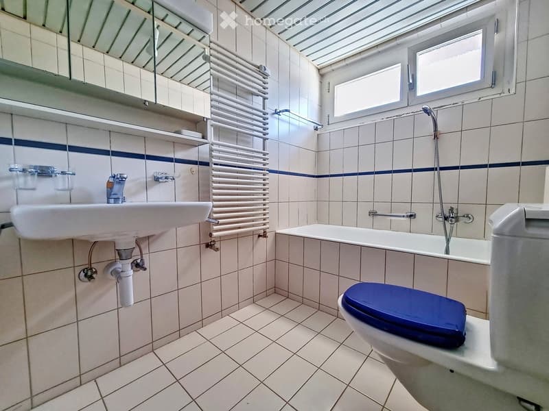 Bathroom with white tiles, a sink, a toilet with a blue seat, a bathtub, a radiator, and a mirror above the sink.
