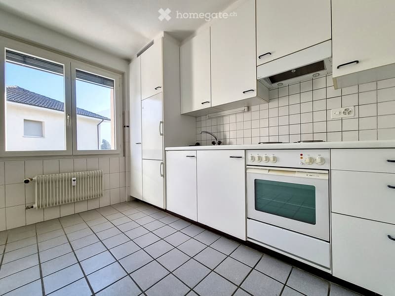 Modern kitchen with white cabinets, tiled walls, and a built-in oven. Features a tiled floor, radiator, and windows offering a view of the roof.