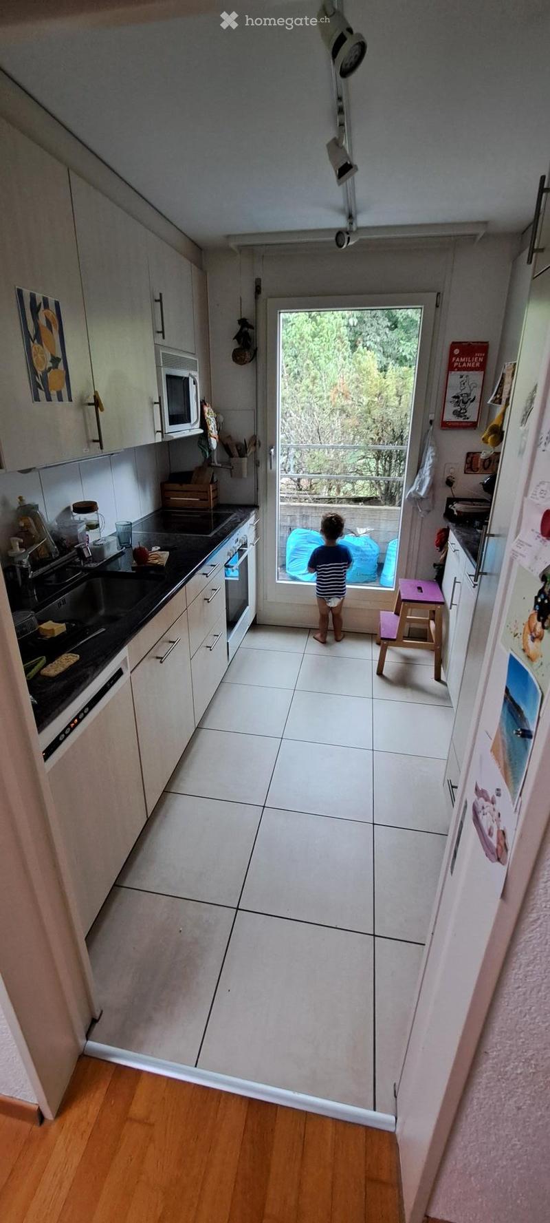 Modern kitchen with white cabinets, black countertops, and a tiled floor. A child stands near a sliding glass door leading to a balcony.