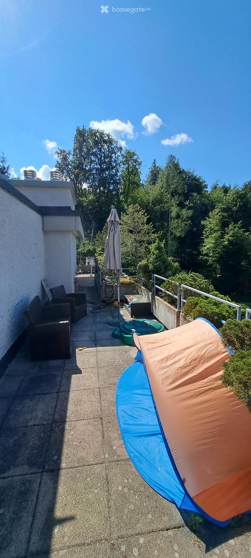 Balcony with a sofa, umbrella, and small pool, surrounded by greenery and a clear blue sky.