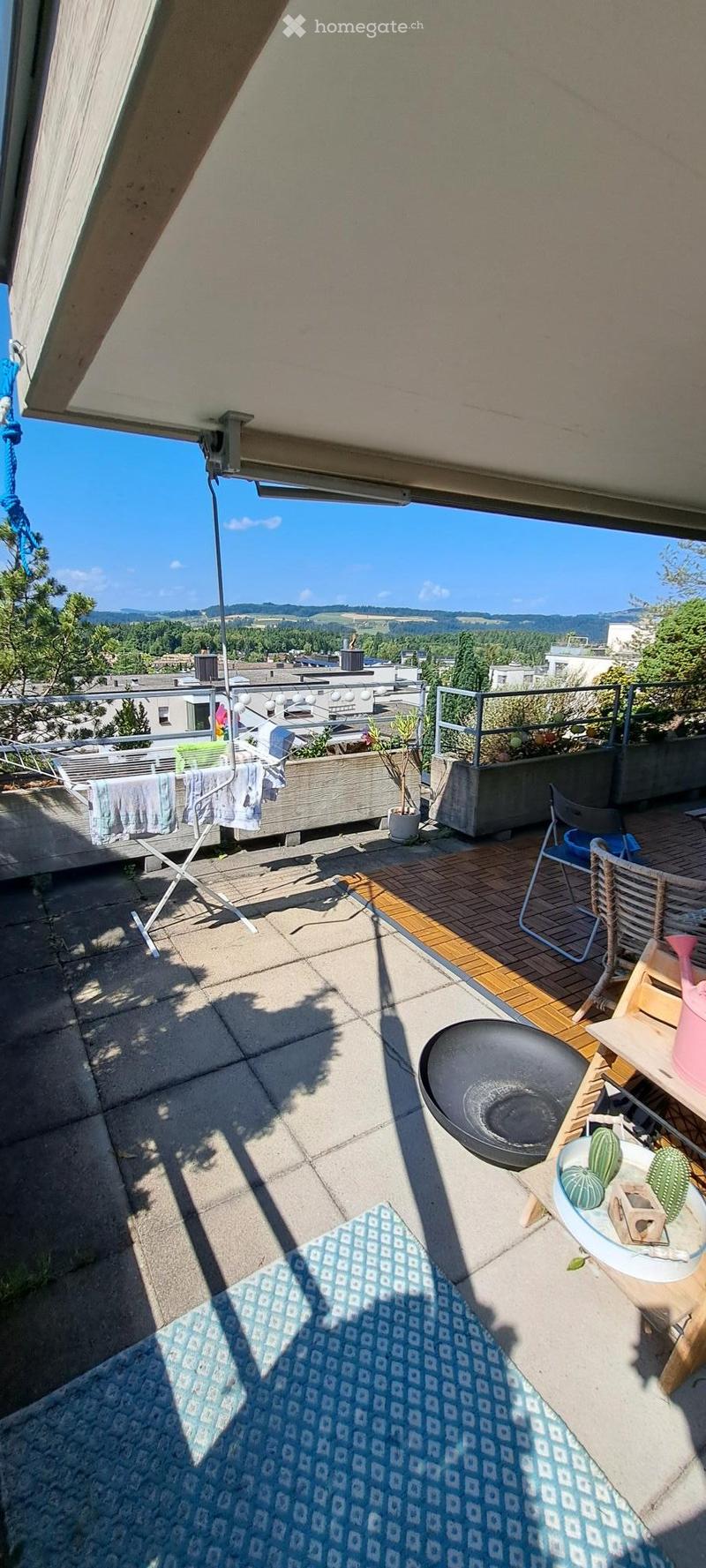 Balcony with wooden decking, concrete floor, outdoor furniture including a table, chairs, and a rug. There is a clothes drying rack, potted plants, and a scenic view of the city and surrounding landscape.