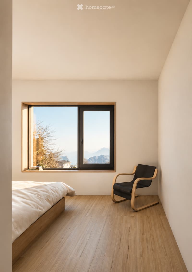Minimalist bedroom with light wooden flooring, a simple bed with white bedding, a wooden-framed chair, and a large window offering a scenic mountain view.
