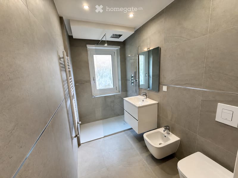 Modern bathroom with beige tiled walls, a white sink with a mirror, a shower area with glass enclosure, a bidet, and a toilet.