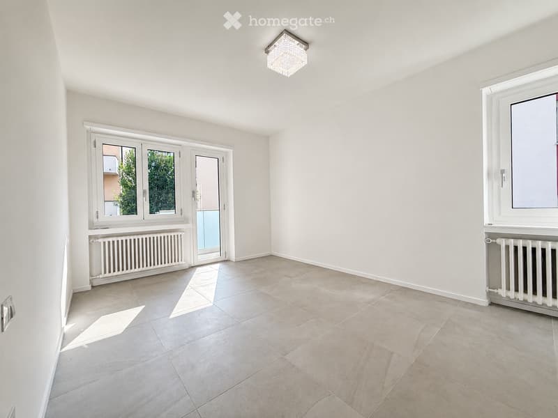 Empty room with light-colored tiled flooring, white walls, and large windows. Features a radiator, a door leading outside, and a chandelier on the ceiling.