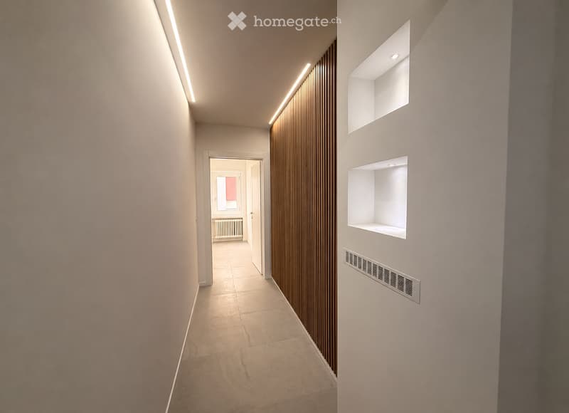 Modern hallway with light-colored tiled flooring, white walls, built-in shelving, and recessed lighting. Features a wooden panel accent wall and a visible doorway leading to another room.