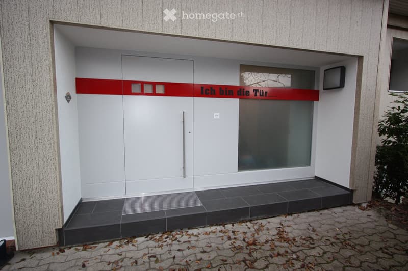 Modern building entrance with a white double door, red accent strip, and small window to the right. The entrance has a small step and is surrounded by a textured concrete wall and a paved area with scattered leaves.
