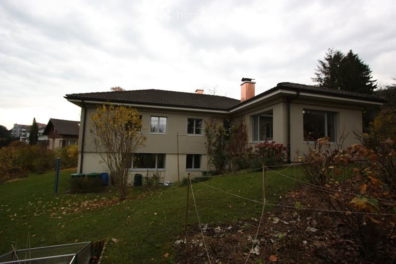Two-story house with beige siding, multiple windows, and a chimney. The front yard has grass, a clothesline, and some plants.