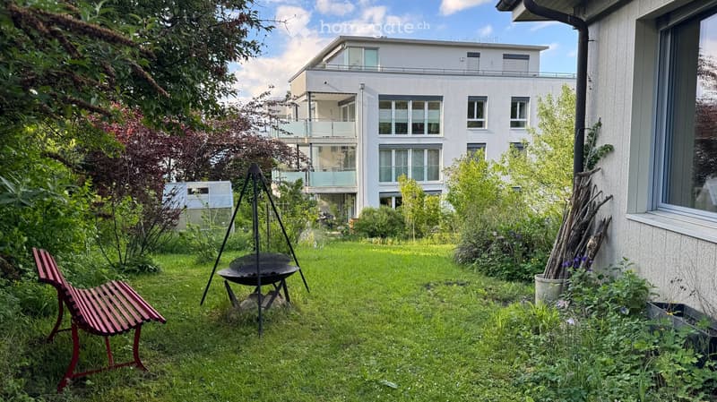 Garden with a red bench, a round metal grill, and lush greenery, with a modern multi-story building in the background.