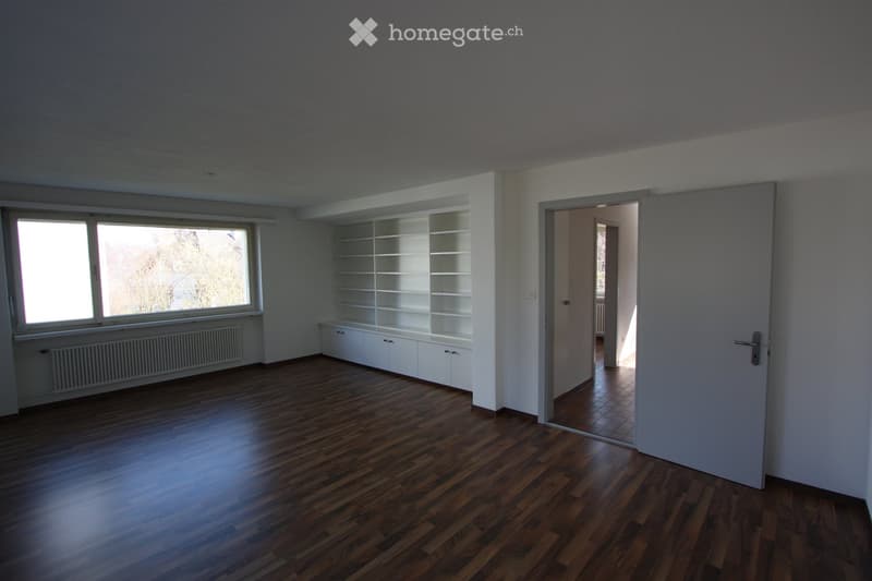 Empty room with wooden flooring, white built-in shelving unit, large window with radiator below, and an open doorway leading to another room.
