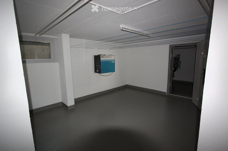 Empty room with white walls, black floor, exposed ceiling pipes, and a small window. A blue electrical panel is mounted on one wall, and an open doorway leads to another space.