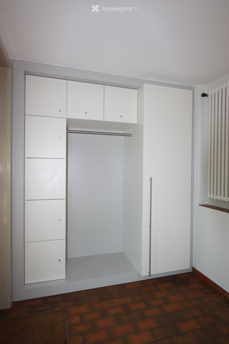 Empty room with white built-in wardrobes, a clothes rail, and a radiator. The floor is made of red tiles.