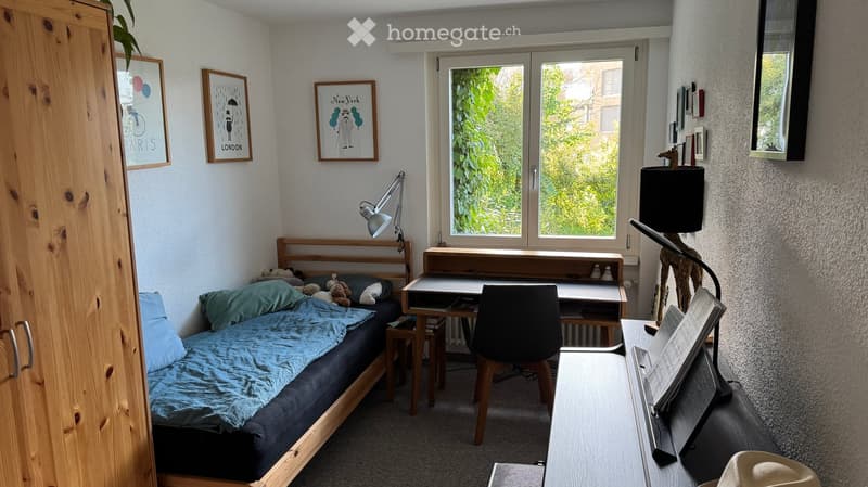 Cozy bedroom with a wooden bed, desk, chair, and a wardrobe. Features a large window with a view of greenery, a lamp on the desk, and decorative picture frames on the wall.