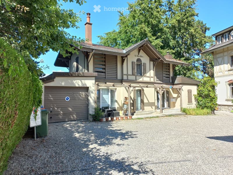 Two-story house with a garage, surrounded by greenery and a gravel driveway.