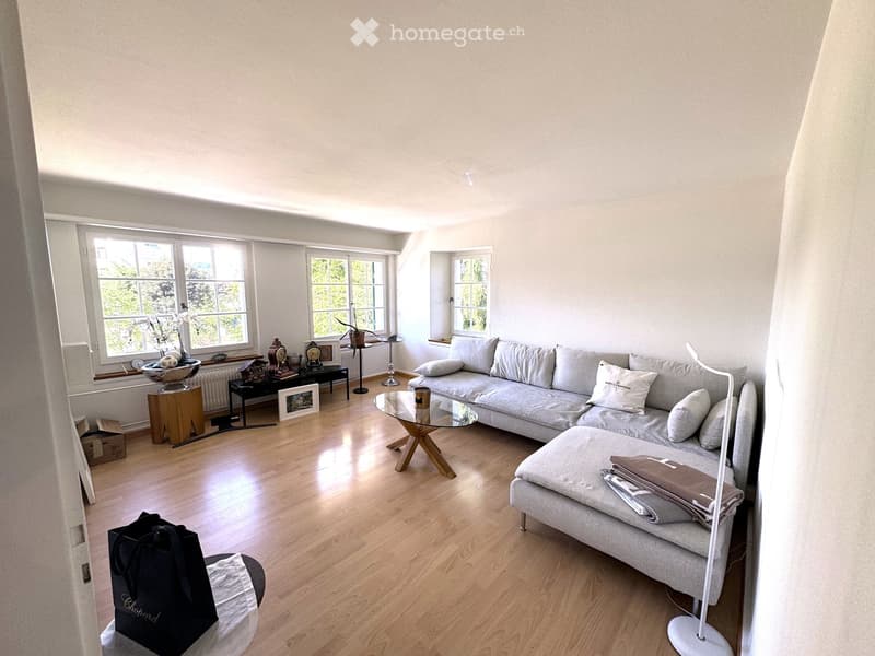 Bright living room with light-colored walls, wooden flooring, large windows, grey sectional sofa, glass coffee table, ottoman, side table, decorative items, and a handbag on the floor.