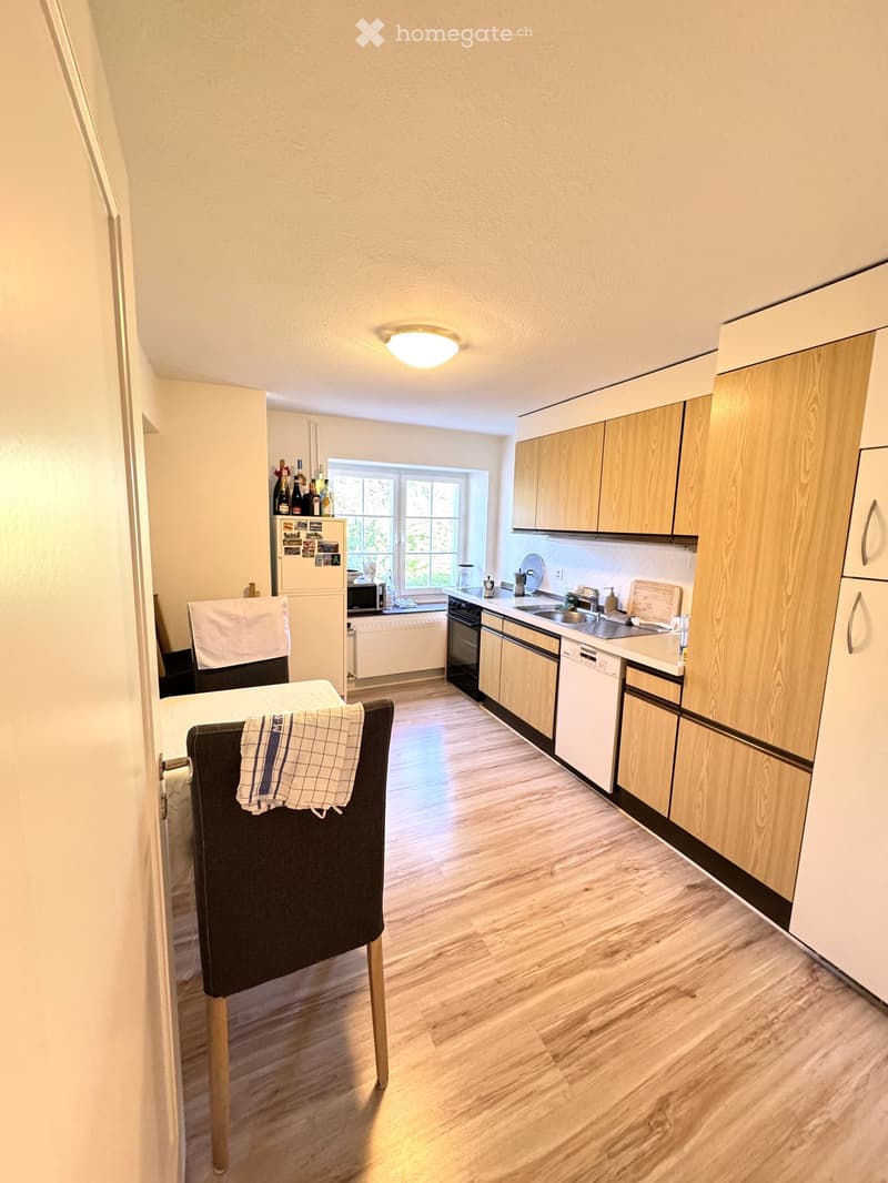 A modern kitchen with light wooden cabinets, stainless steel appliances, a dining area with a table and chairs, and a window letting in natural light.