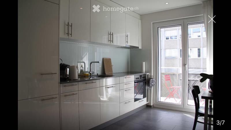 Modern kitchen with white cabinets, stainless steel appliances, and a large window overlooking a balcony area with outdoor furniture.