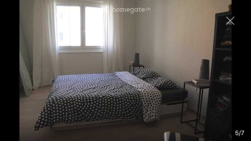 A cozy bedroom with a double bed covered in a black and white polka dot duvet, a nightstand with a lamp, and a tall shelving unit with books and decorative items.
