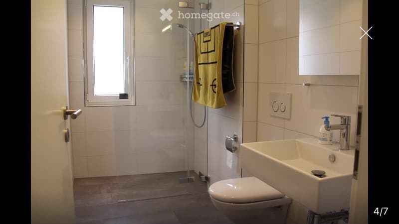 Modern bathroom with white tiles, a glass-enclosed shower, a toilet, a sink with a modern faucet, and a yellow towel hanging on the wall.