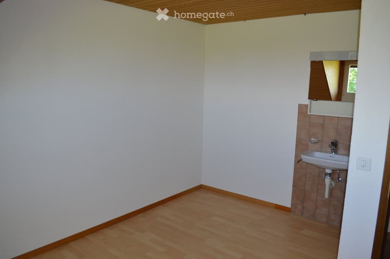 Empty room with light wooden flooring, white walls, and a small sink area with a mirror and tiled backsplash.