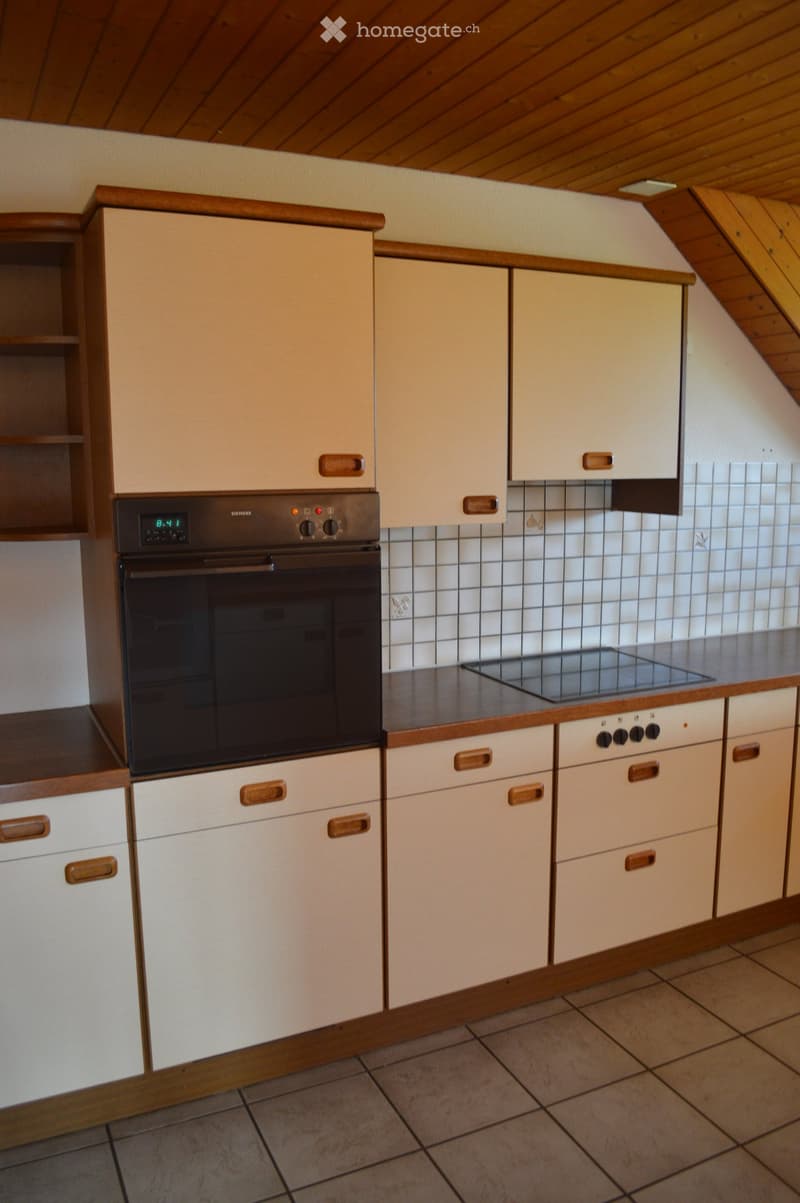 A modern kitchen with white cabinets, wooden trim, and a built-in oven and stove. The countertops are dark, and the floor is tiled.