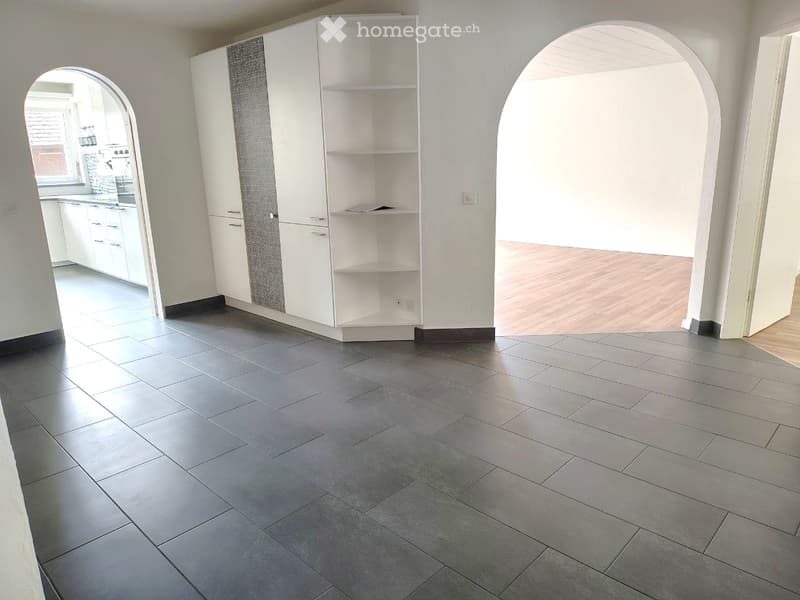 Empty room with grey tiled floor, white walls, and white built-in shelving unit. Adjacent kitchen area visible through an arched doorway.