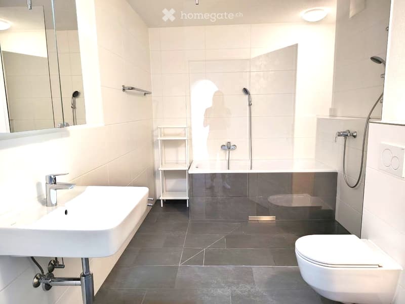 Modern bathroom with white tiles, a sink, toilet, shower, and a small shelf. The shower has a glass enclosure, and the room features sleek chrome fixtures.