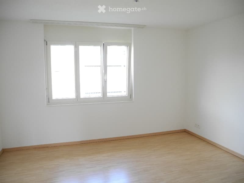 Empty room with light wooden flooring and white walls, featuring a large window with white frames and blinds.