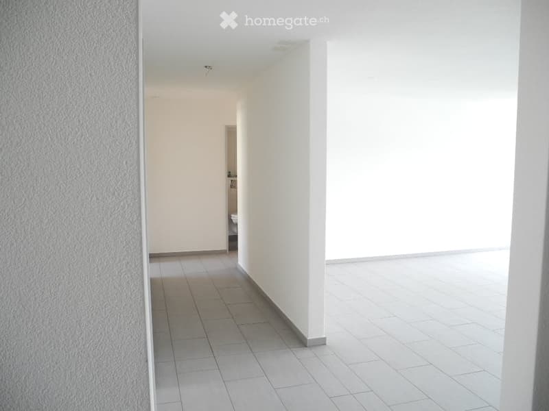 Empty hallway with white tiled flooring and white walls, leading to an open area with large windows.