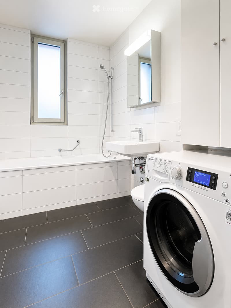 Modern bathroom with white tiles, black tiled floor, bathtub, sink, mirror, shower, and a private washing machine.