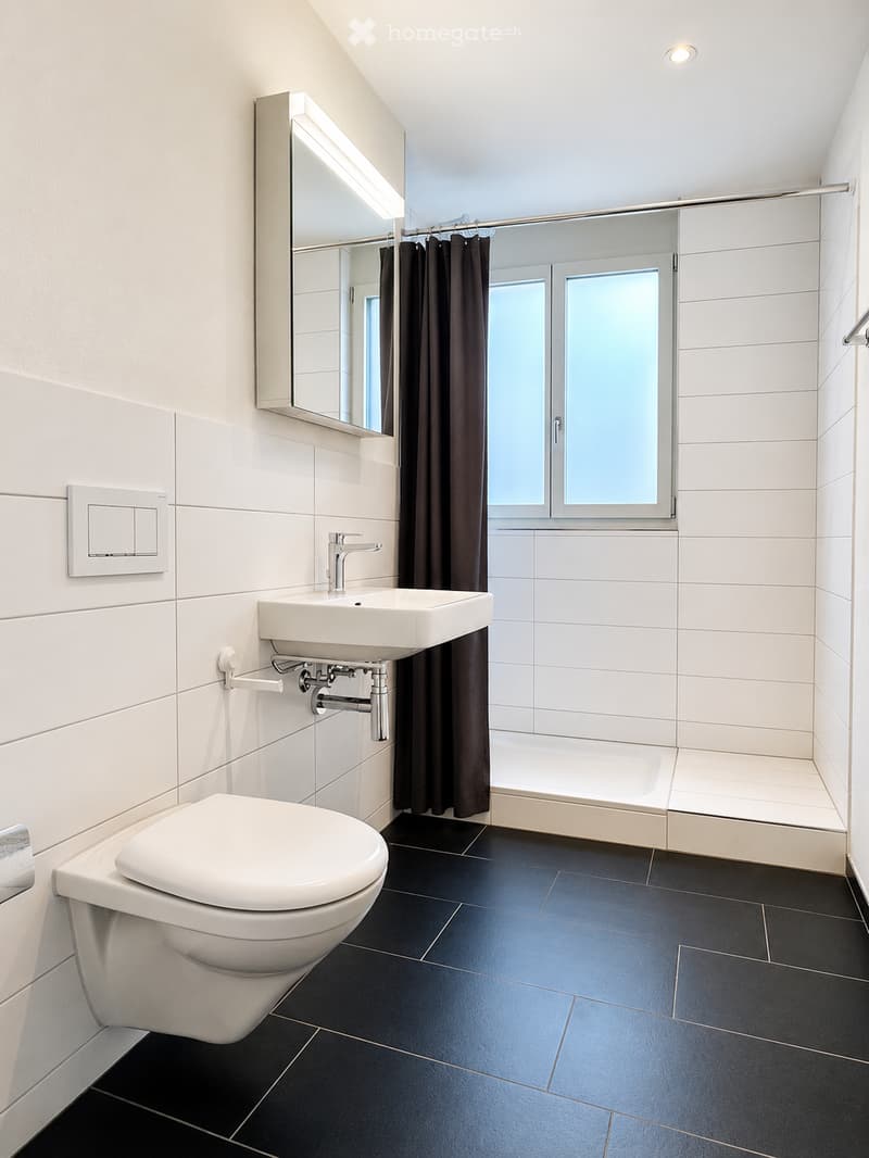 Modern bathroom with white tiles, black floor, toilet, sink, mirror, and shower area with dark curtains.