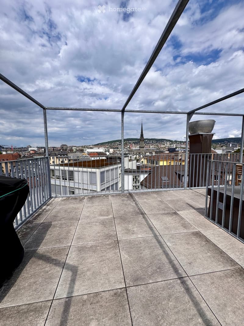 Roof terrace with concrete tiles, metal railing, and a view of the city skyline and a church spire.