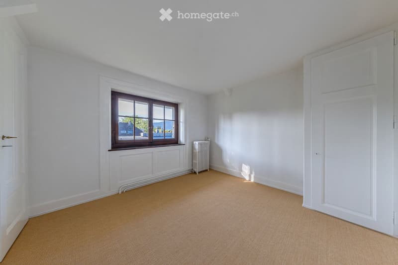 Empty room with white walls, carpeted floor, a window with brown frame, radiator, and white doors.
