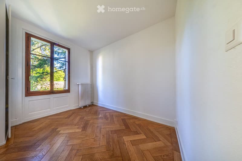 Empty room with white walls, wooden herringbone flooring, and a large window with wooden frame. A radiator is visible below the window.