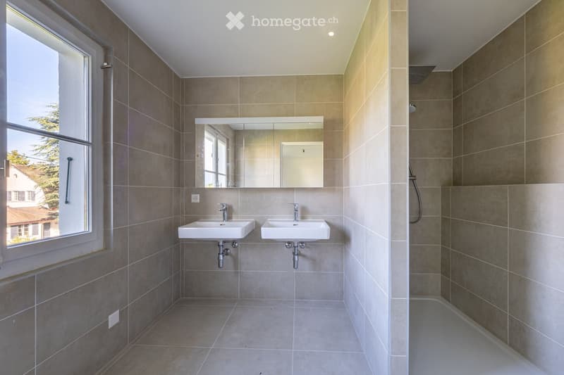 Modern bathroom with two white sinks, a large mirror, and a bathtub, featuring beige tiles and a large window with a view of the outside.