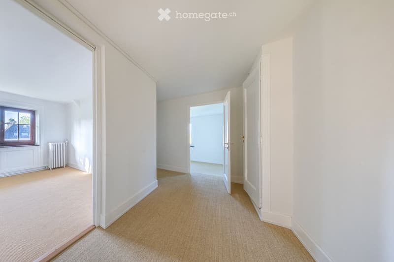 Empty hallway with beige carpet, white walls, and a window with a radiator below it. The hallway leads to another open doorway.