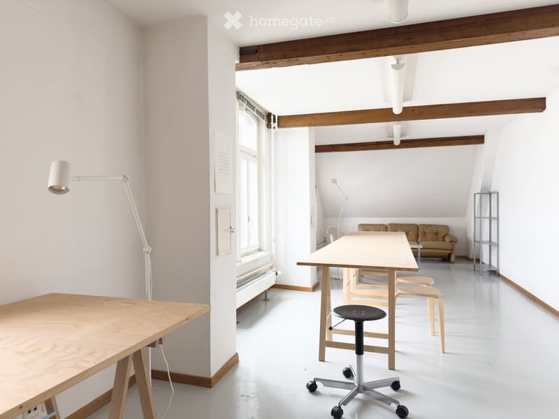 Bright, open room with white walls and wooden beams, featuring a large central table, chairs, a sofa, and a stool.