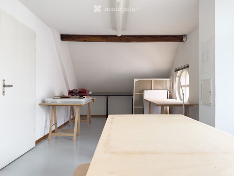 Bright, minimalist room with white walls and exposed wooden beams. Contains a large central table, a smaller table with a stool, a shelf, and a lamp near a window.