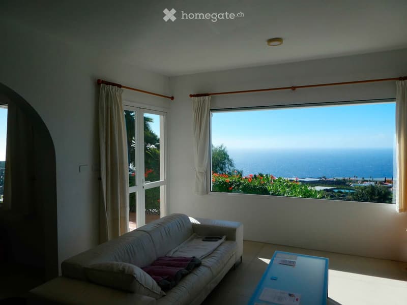 Bright living room with large windows offering a scenic ocean view, featuring a white sofa and a glass coffee table.