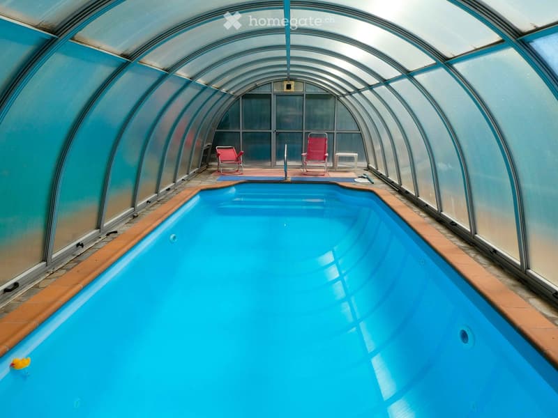 Indoor swimming pool area with blue water, surrounded by glass walls, two red chairs, and a small table.