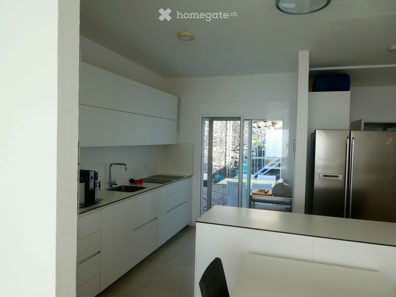 Modern kitchen with white cabinets, stainless steel appliances, and a large island. The space features a sink, faucet, coffee maker, and a refrigerator, with a view to an outdoor area through large glass doors.