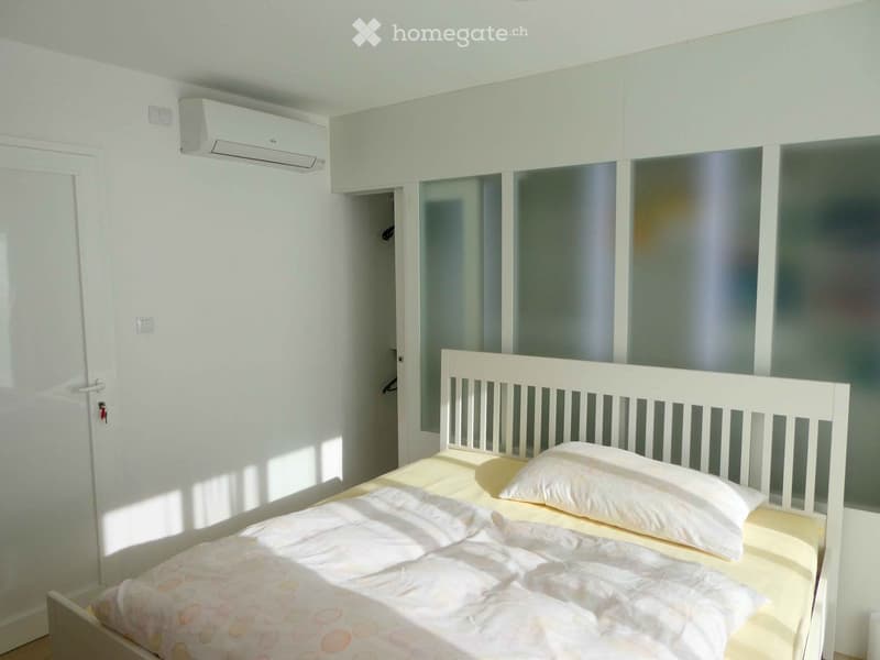 Bright bedroom with a single bed, white headboard, and matching bedding. An air conditioning unit is mounted on the wall, and a closet with mirrored doors is visible in the background.