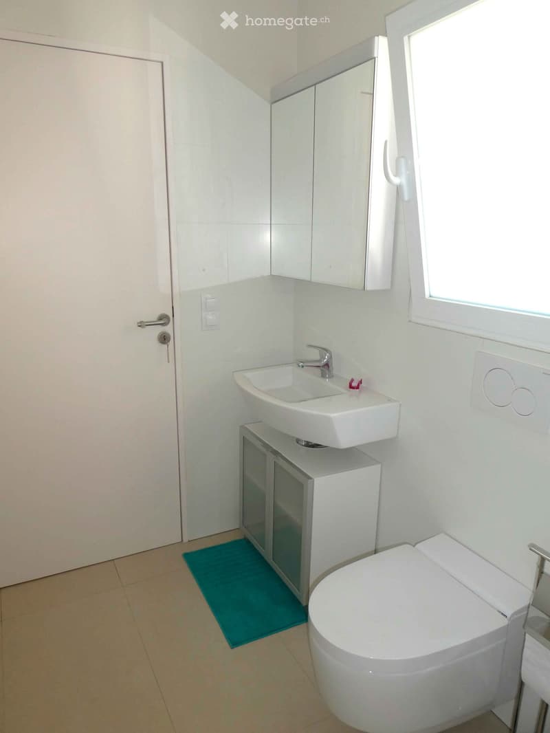 Modern bathroom with white tiles, a sink with a faucet, a toilet, a mirror cabinet, and a blue bathroom mat on the floor.