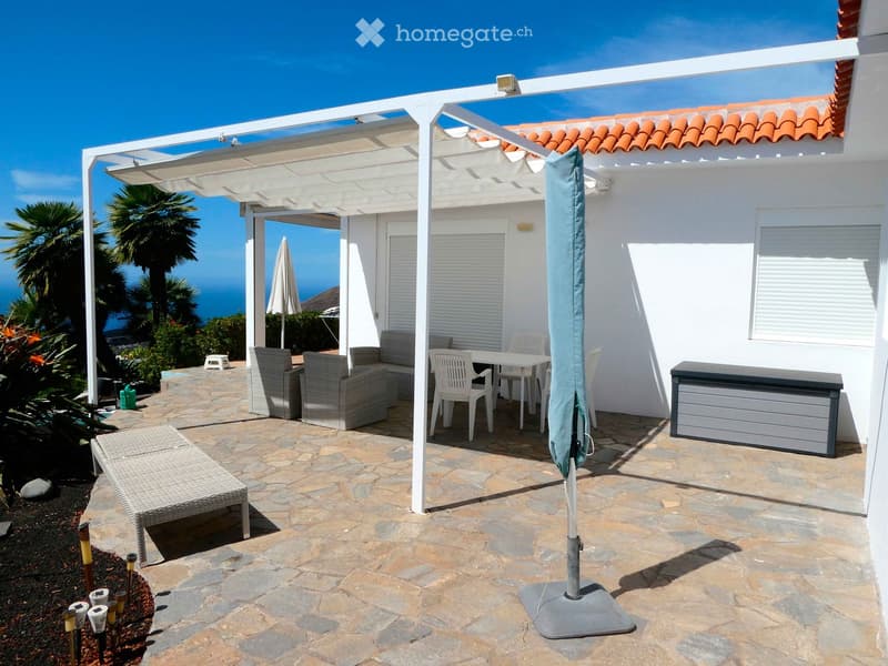 Outdoor patio with stone flooring, white pergola, sofa, table, chairs, and umbrella, overlooking the ocean.