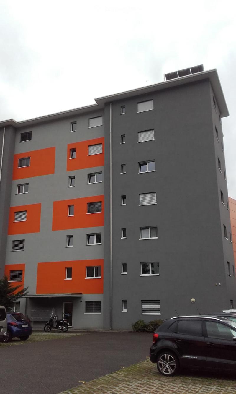 Modern multi-story building with grey and orange facade, multiple windows, and a small entrance area with a parked scooter and cars nearby.